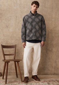 Tikkidega jakk geomeetrilise mustriga musta ja kreemika tooniga, lukuga ja kõrge kraega. Kanditud valgete laiade pükstega ja pruunide kingadega.