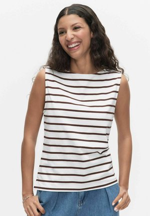 Vrouw met lang krullend haar lacht en draagt een mouwloos wit topje met horizontale bruine strepen en een blauwe spijkerbroek tegen een effen achtergrond.