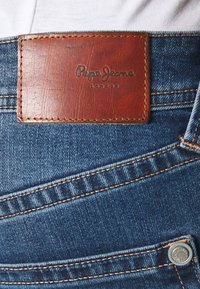 Denim jeans feature a textured blue fabric with a brown leather patch embossed "Pepe Jeans London" and contrasting yellow stitching accents.