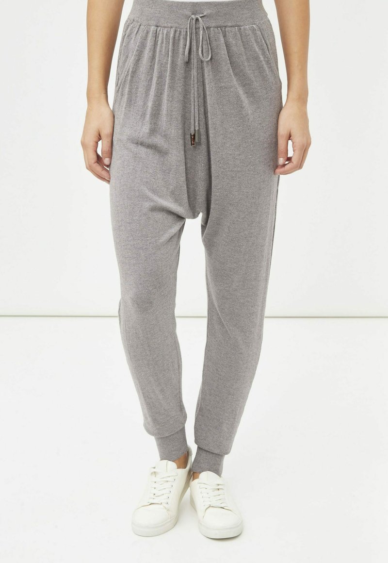 Pantalones de jogging gris de punto con cintura ajustable, puños cónicos y bolsillos laterales, combinados con zapatillas blancas. La tela parece suave y cómoda.