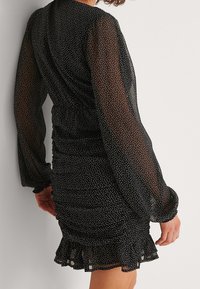 Vestido negro con lunares blancos, mangas largas transparentes, detalles fruncidos y un dobladillo con volantes. La tela parece ligera y texturada.
