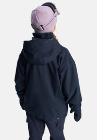Person wearing a black hooded jacket, black gloves, navy pants, and a pink knit hat with ski goggles resting on top, viewed from behind.