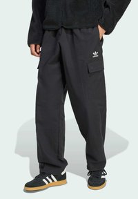 Pantalones cargo negros de corte relajado, con bolsillos laterales y un logo de Adidas. Combinados con zapatillas negras con rayas blancas y suela de goma.
