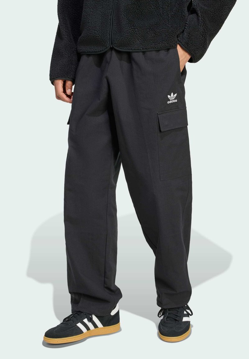 Pantalones cargo negros de corte relajado, con bolsillos laterales y un logo de Adidas. Combinados con zapatillas negras con rayas blancas y suela de goma.