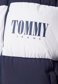 Närbild av en navy- och vit västjacka med "TOMMY JEANS"-logotyp tryckt på bröstet ovanför en sidogömma.