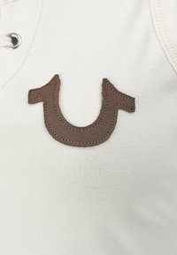 Gros plan sur un tissu blanc avec un emblème marron en forme de fer à cheval cousu et un œillet métallique en haut à gauche.