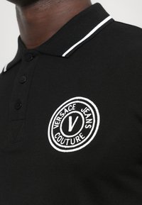 Cămașă polo neagră cu o deschidere la gât cu trei butoane, dungi albe la guler și un logo circular brodat care prezintă "Versace Jeans Couture."