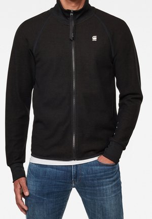 Mann trägt eine schwarze, zum Zuziehen geeignete Jacke über einem weißen Hemd mit blauen Jeans, eine Hand in der Tasche, vor einem schlichten hellen Hintergrund.