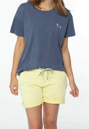 Femme portant un t-shirt bleu à manches courtes et un short jaune clair à ourlet roulé avec une ceinture à cordon florale, debout devant un fond blanc.