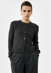 Mujer con peinado trenzado oscuro que lleva pendientes de aro plateados, un cárdigan abotonado de punto cable gris oscuro y pantalones gris oscuro con pliegues.