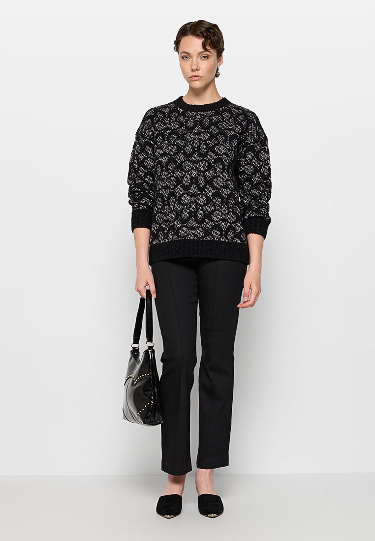 Vrouw wearing een zwart patroontrui, zwarte strakke broek, zwarte flats, houdend een zwarte handtas met gouden studs, staand tegen een witte achtergrond.