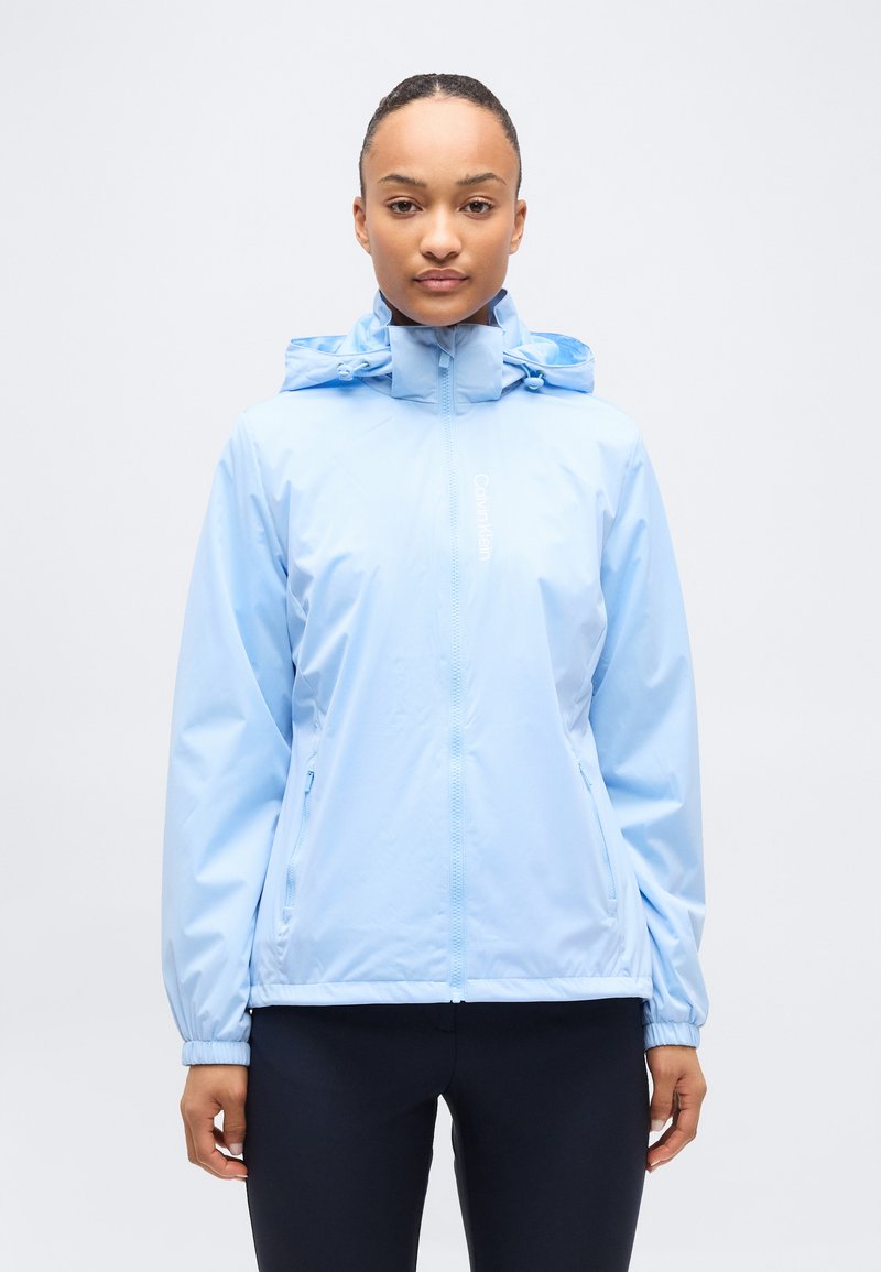 Γυναίκα που φοράει ανοιχτό γαλάζιο τζάκετ βροχής με κουκούλα, φερμουάρ μπροστά και πλαϊνές τσέπες, που στέκεται μπροστά σε ένα απλό ανοιχτό γκρι φόντο.