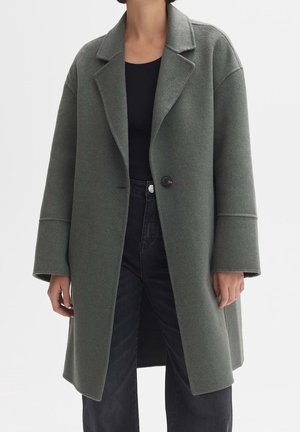Femme portant un long manteau gris en laine oversize avec de larges revers, un bouton unique, un haut noir et un pantalon noir taille haute.