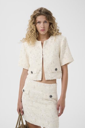 Kvinde med krøllet hår iført en cremefarvet jakke med korte ærmer og pailletter samt matchende nederdel, der holder en beige håndtaske foran en ensfarvet baggrund.