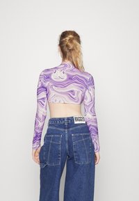 Camisola cropped de manga comprida em roxo marmoreado combinada com jeans de ganga azul de cintura alta. Os jeans apresentam uma etiqueta de marca visível "RAGGED."