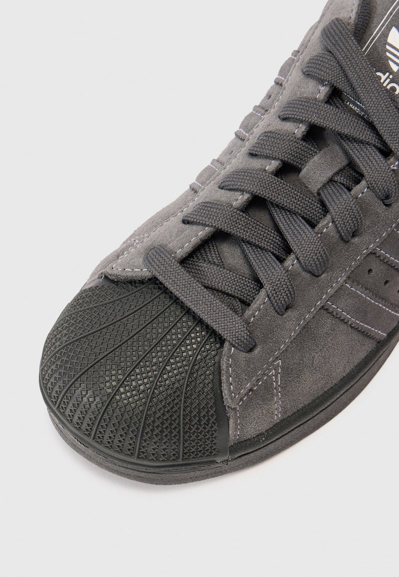 Baskets en daim gris avec un embout noir texturé, des lacets plats noirs et des détails de couture contrastants sur la tige.