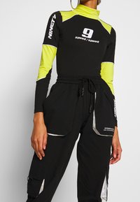 Zwarte lange mouwen bodysuit met gele accenten en witte grafische tekst; gecombineerd met zwarte cargobroek met reflecterende details en zakken.