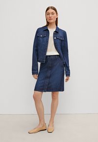 Veste en denim foncé avec deux poches poitrine, associée à une jupe en denim assortie. La tenue comporte un haut de couleur claire et des ballerines beiges.
