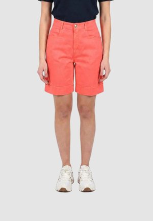Persona che indossa pantaloncini corallo fino al ginocchio, top nero e scarpe da ginnastica bianche, in piedi davanti a uno sfondo chiaro uniforme.