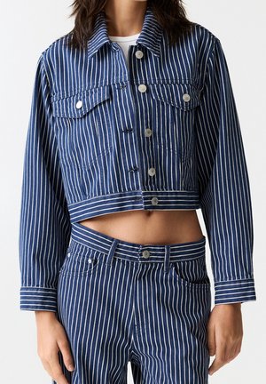 Woman wearing a cropped blue denim jacket and matching high-waisted pants with white vertical stripes and silver buttons.