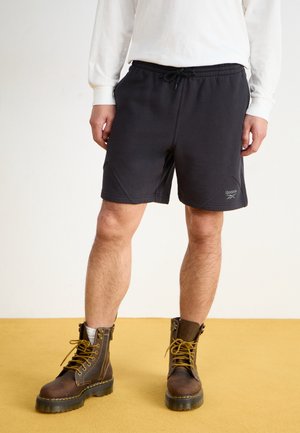 Mann, der schwarze Reebok-Shorts, ein weißes Langarmshirt und braune Schnürstiefel trägt und auf einem gelben Boden steht.