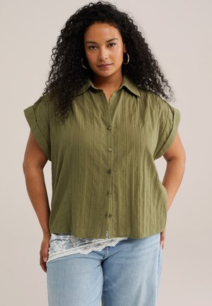 Göndör hajú nő arany karika fülbevalóval, olívazöld rövid ujjú gombos ingben és világoskék farmerben, amely csipkedíszítéssel rendelkezik.