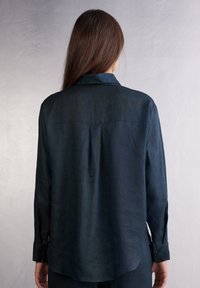 Camicia a maniche lunghe in tessuto leggero di un blu navy scuro. Presenta un colletto classico, polsini con bottone e una spallina sulla schiena.