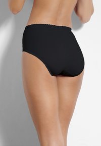 Donna che indossa delle mutande nere a vita alta, vista da dietro, in piedi contro un muro bianco e grigio.