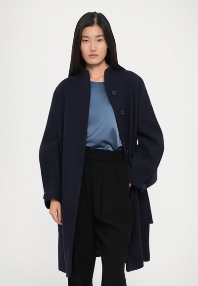 Femeie cu păr lung și dark, purtând un palton oversized bleumarin cu nasturi, un top din satin albastru și pantaloni negri cu talie înaltă, având o mână în buzunar.