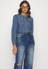 Camisa de деним com colarinho em babados, azul, mangas longas e fecho de botão. Usada com jeans de деним desgastados com um remendo estampado.