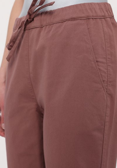 Brązowe, lekkie, zwężane spodnie z elastycznym paskiem, kieszeniami bocznymi i gładką fakturą. Posiadają subtelny połysk i staranne szwy.
