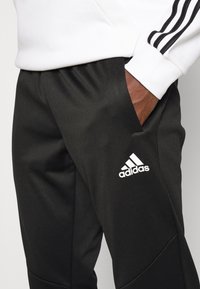 Černé sportovní kalhoty s hladkým povrchem, bílým logem Adidas a bočními kapsami. Pas je elastický pro pohodlí.