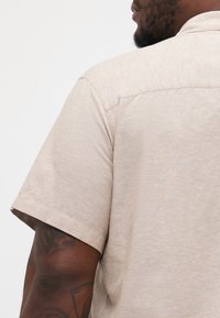 Camicetta beige a maniche corte con una texture sottile, caratterizzata da un colletto rilassato e dettagli sulla cucitura della spalla. Tatuaggio prominente visibile sul braccio.