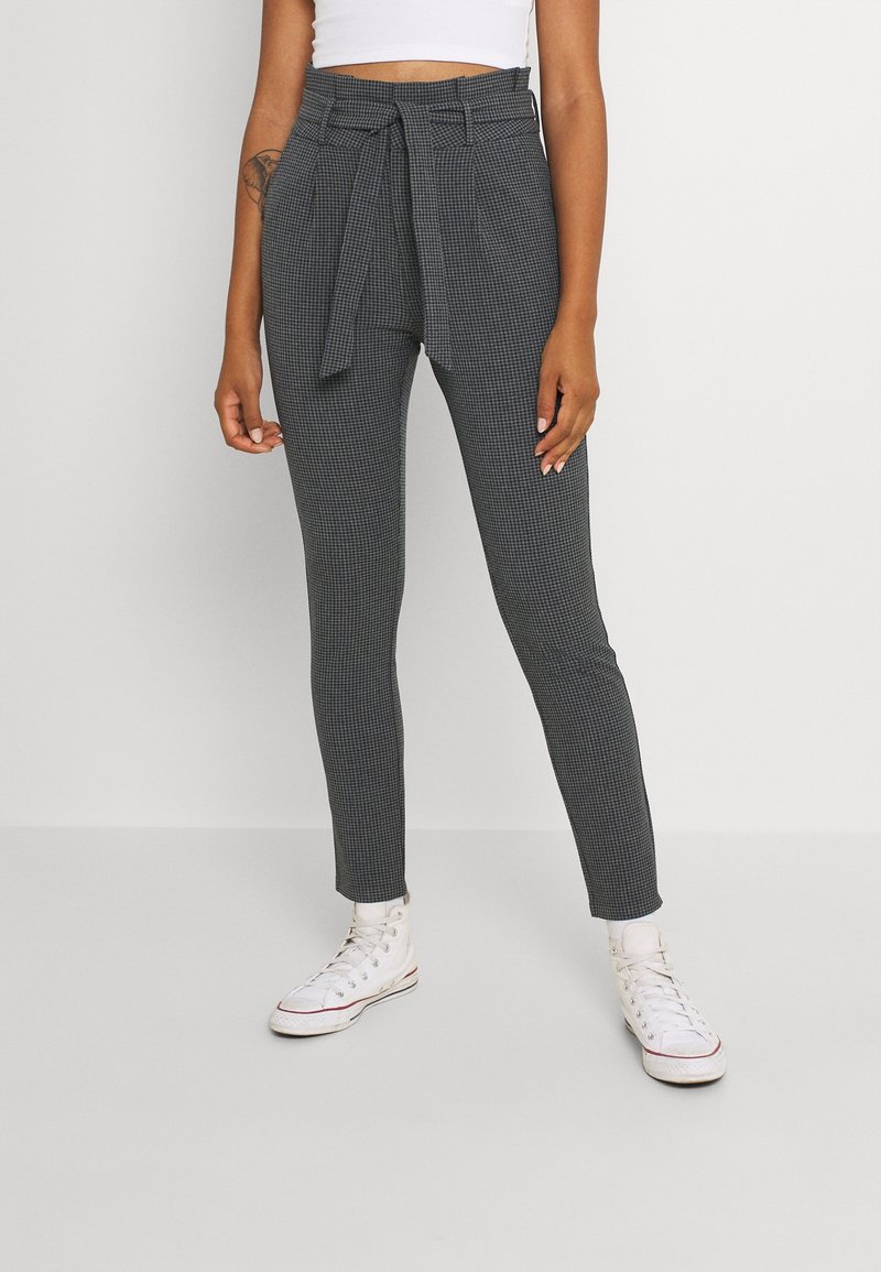 Femme portant un pantalon à carreaux gris taille haute avec une ceinture en tissu, un top court blanc et des baskets blanches montantes sur un fond uni.