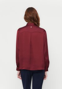 Camicia di raso borgogna con maniche lunghe, polsini con bottoni e piega posteriore. Presenta un piccolo logo circolare sul colletto.