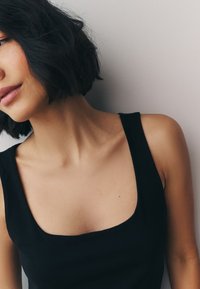 Čierne tielko bez rukávov so štvorcovým výstrihom; hladká textúra a priliehavý strih. Pozadie je svetlosivé, so zameraním na odev.