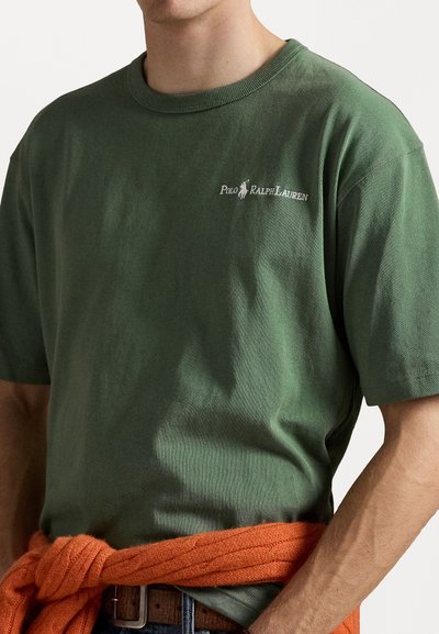 Green cotton T-shirt with crew neckline and short sleeves, featuring a white Polo Ralph Lauren logo on the left chest, worn with an orange sweater tied at the waist.