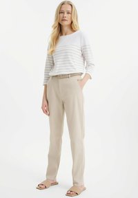 Pasiasta biała i beżowa koszula z długim rękawem w połączeniu z beżowymi spodniami, z plecionym paskiem i płaskimi sandałami, na jednolitym tle.