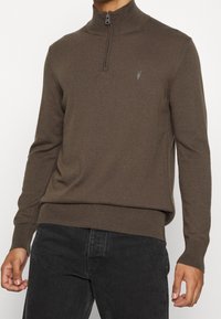 Hnědý svetr na zip s žebrovanými manžetami a lemem. Vyrobený z měkkého materiálu, s malým logem na hrudi a vysokým límcem.