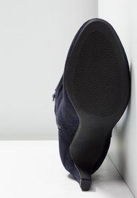 Botte à talon haut en daim bleu marine à semelle noire reposant de côté contre un angle blanc de mur et de sol.