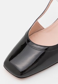 Chaussure slingback en cuir verni noir avec bout carré et doublure intérieure beige, présentée en gros plan sur fond blanc.