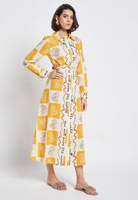 Long, collared dress in white and yellow with floral and text patterns. Fabric appears lightweight; features a cinched waist and button details.