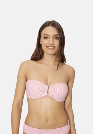 Joven con cabello largo y castaño que lleva un top de bikini sin tirantes a rayas rosas y blancas, sonriendo frente a un fondo claro y liso.