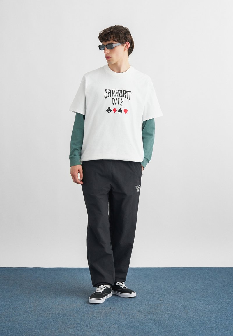 Junger Mann mit schwarzen Sonnenbrillen, weißem T-Shirt mit Grafik über grünen Langarmärmeln, schwarzen Hosen und schwarzen Turnschuhen steht auf blauem Teppich.