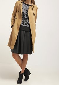 Jasnobeżowy płaszcz trench z podwójnym zapięciem z przodu i naramiennikami, w połączeniu z czarnym swetrem z kwiatowym wzorem i czarną skórzaną spódnicą.