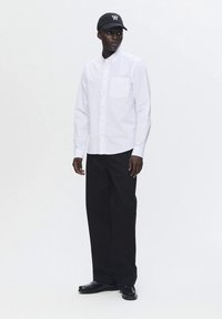 Férfi fehér gombos inget, fekete bő szárú nadrágot, fekete mokaszint és fekete baseball sapkát visel, amelyen fehér logó található.