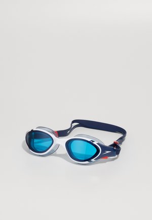 Lunettes de protection avec une monture blanche, des verres teintés en bleu et une sangle marine. Les caractéristiques incluent une sangle ajustable et des détails texturés sur les côtés pour le confort.