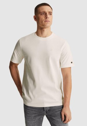 Άνδρας που φοράει απλό λευκό κοντομάνικο T-shirt και γκρι τζιν, στέκεται με το ένα χέρι στην τσέπη, κοιτάζοντας στο πλάι.