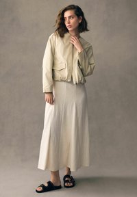 Beezhiga lukuga zip-up bomber jakk, millel on kaks esikot, koos tekstuurse valge pikkade seelikuga ja mustade libisevate sandaalidega, millel on kuldsed aktsendid.