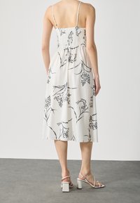 Femme portant une robe midi blanche avec des imprimés floraux noirs et de fines bretelles, associée à des sandales à talons blancs, se tenant sur un sol gris.
